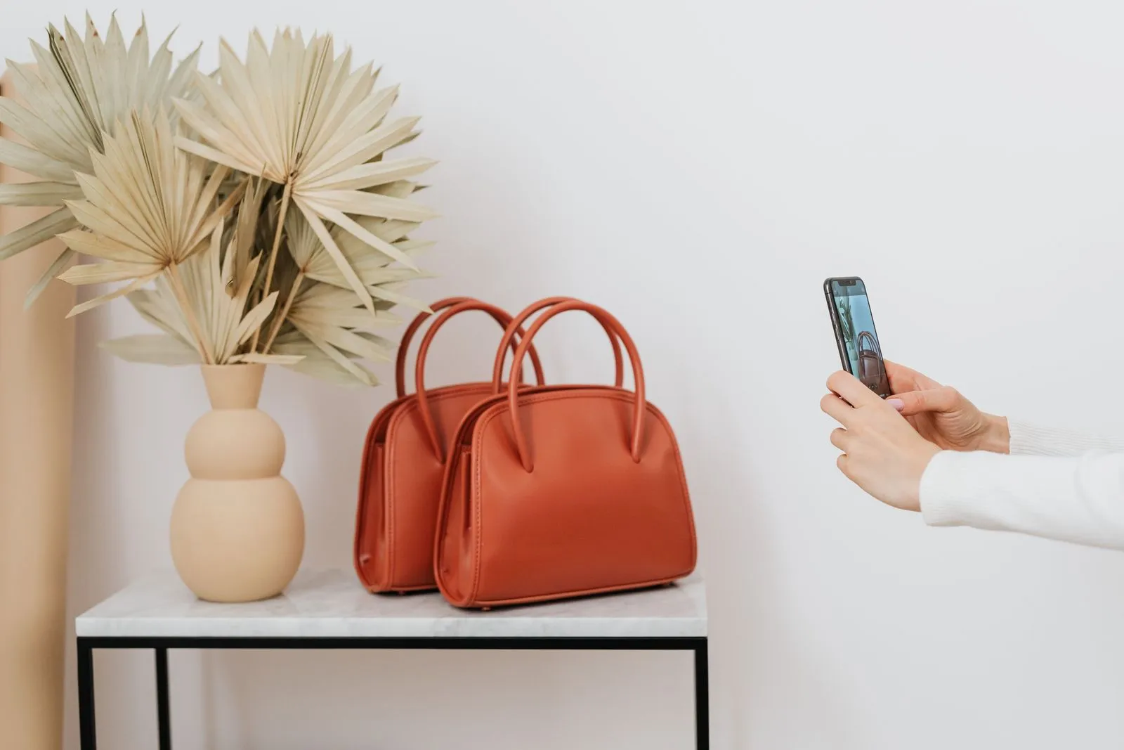 Eine Hand fotografiert mit dem Smartphone zwei rotbraune Ledertaschen auf einem Beistelltisch