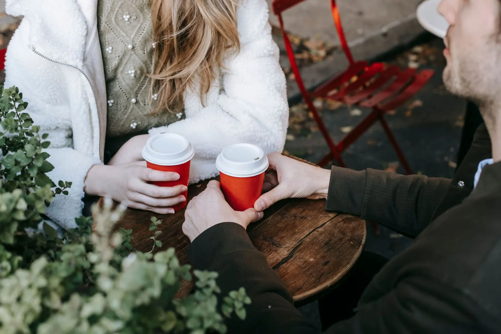 Two people sit at a small wooden table in a sidewalk cafe holding red coffee cups