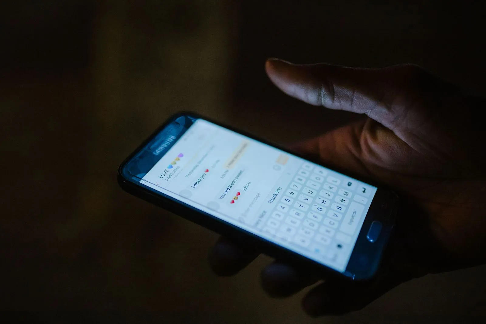 A hand holds a smartphone in the dark, showing a messaging app chat full of heart emojis