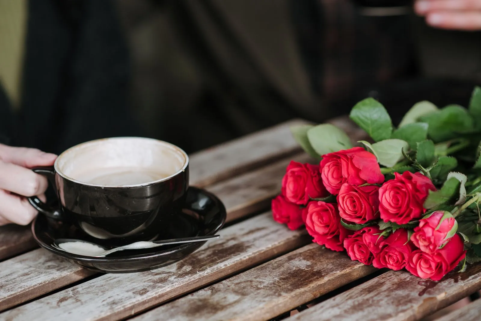 Eine Hand hält eine Tasse Cappuccino auf einem Holztisch, daneben liegt ein Strauß roter Rosen