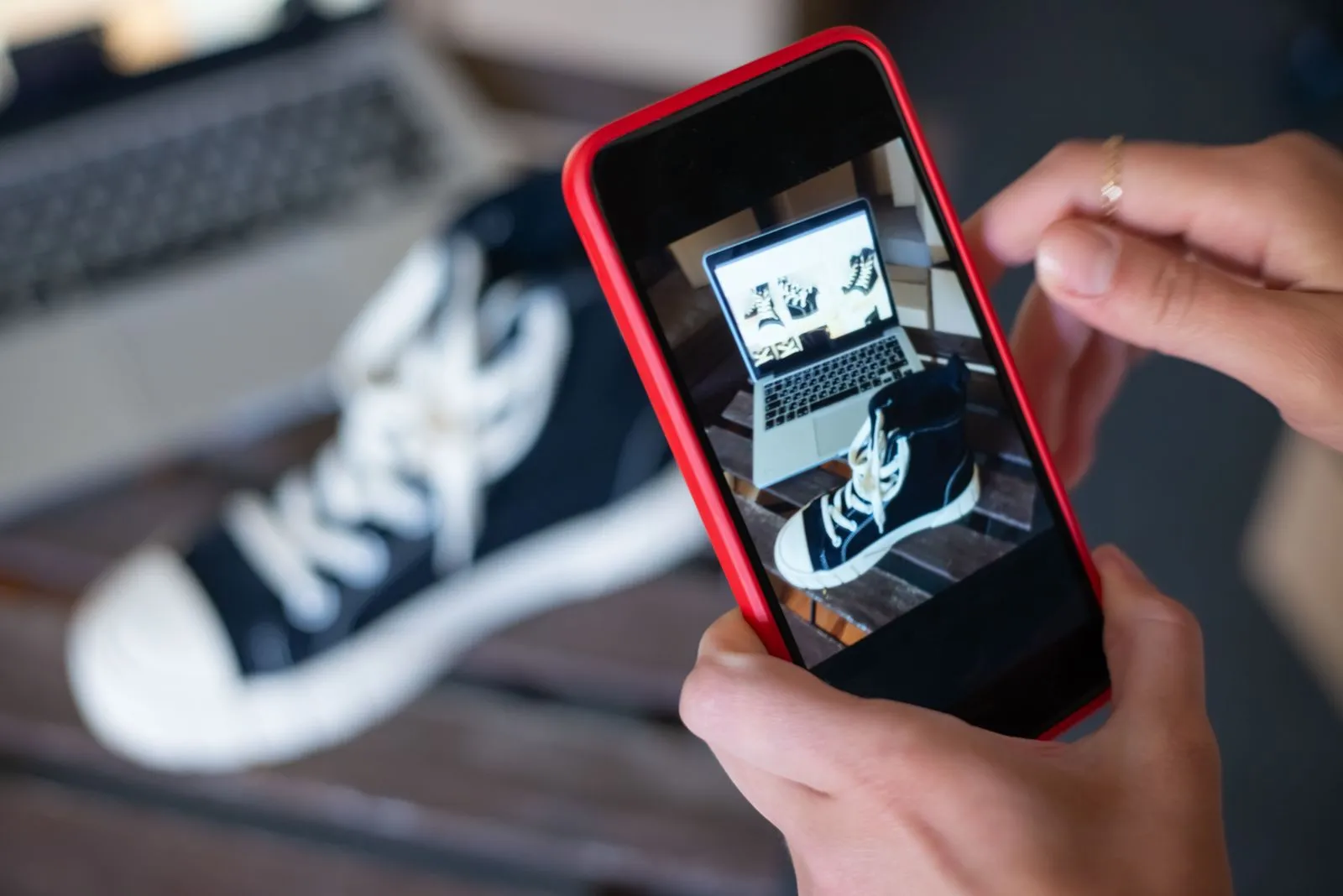 Hände fotografieren einen schwarzen Sneaker mit dem Smartphone, im Hintergrund ein Laptop
