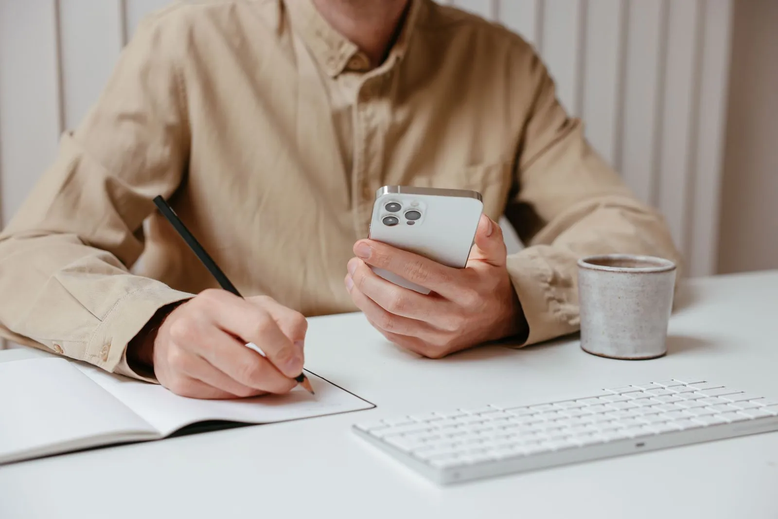 Eine Person schreibt mit Bleistift in ein Notizbuch und hält ein Smartphone, daneben stehen Tastatur und Tasse