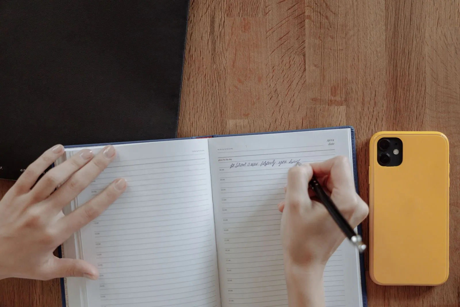 Hands writing in a notebook next to a yellow smartphone on a wooden table