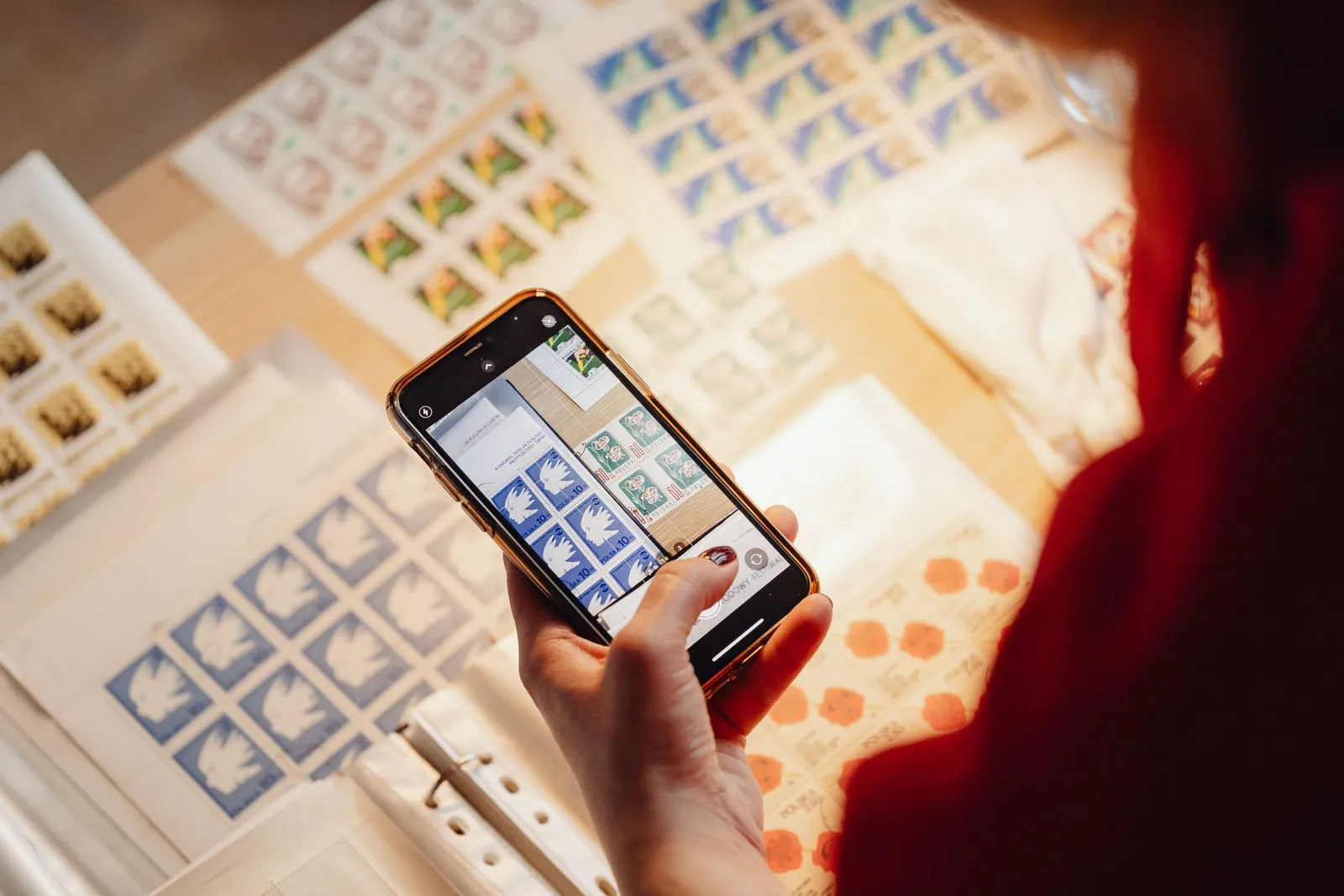 Eine Hand fotografiert mit dem Smartphone Briefmarkenbögen auf einem Tisch, der Bildschirm zeigt die Kamera-App