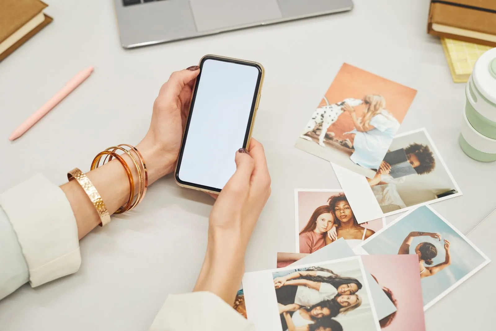 Hände halten ein Smartphone über einem Tisch mit ausgedruckten Fotos, daneben ein Laptop