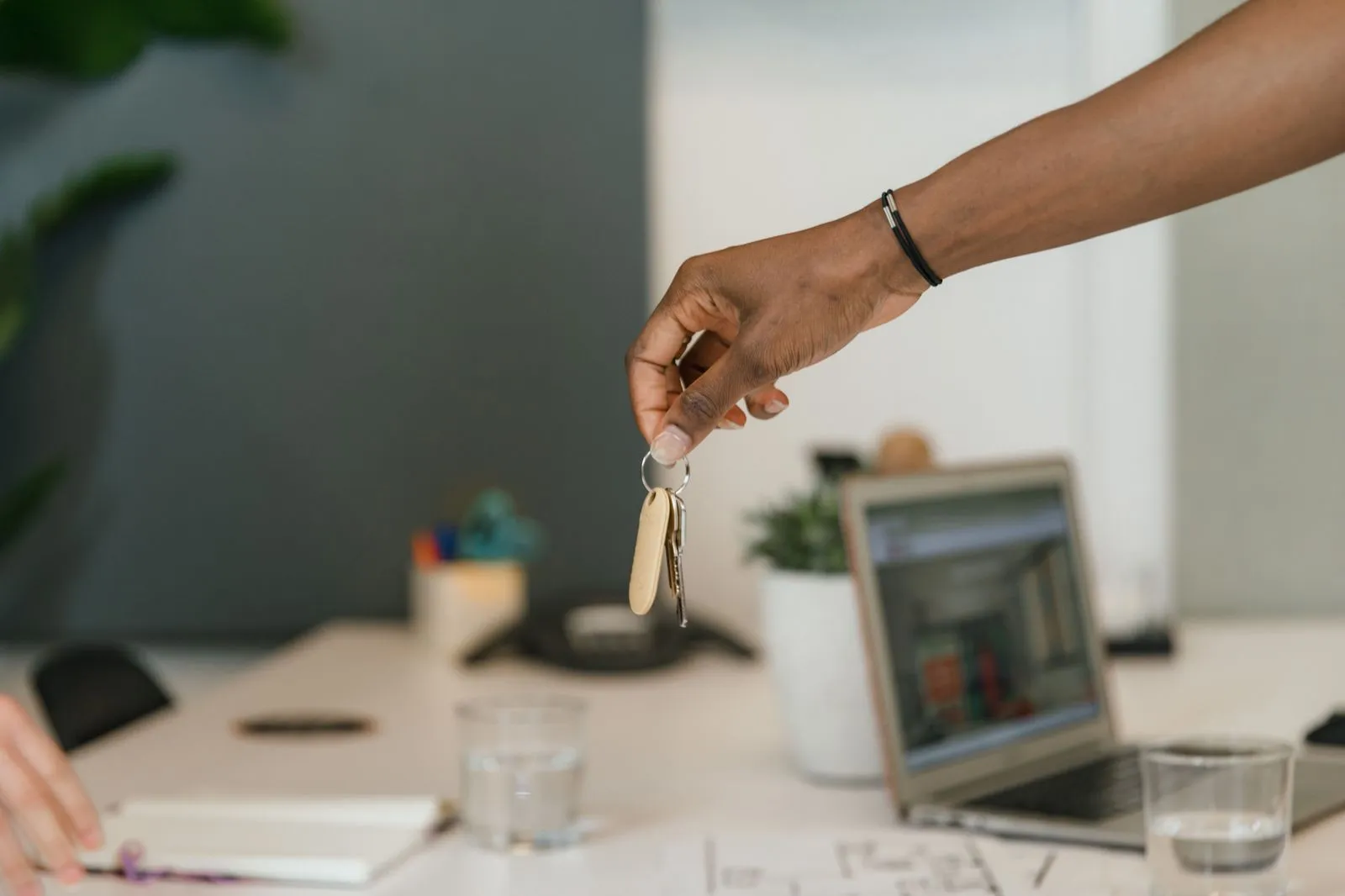 A hand holds a set of keys above a desk with a laptop and a floor plan