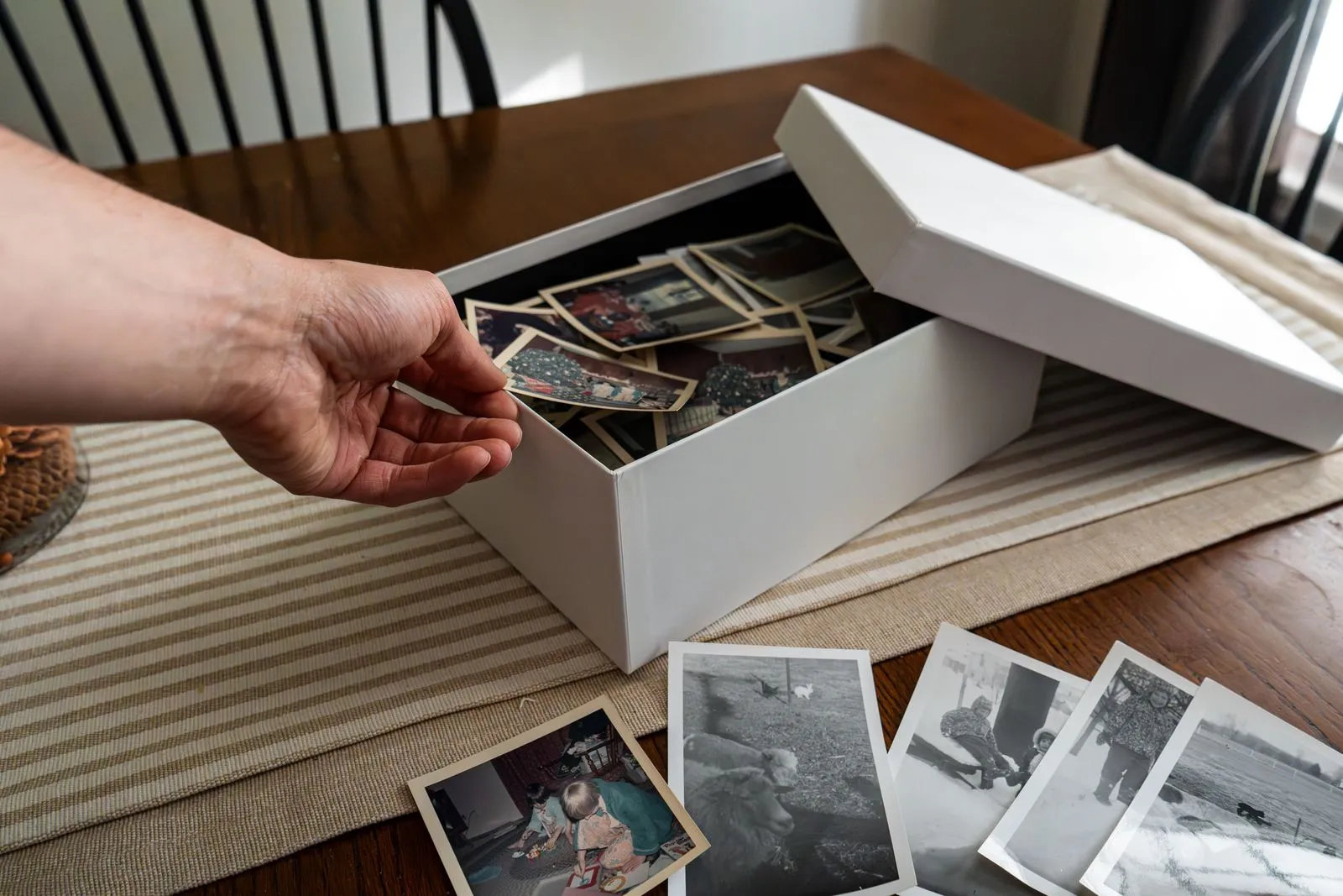 Hand greift in eine Schachtel mit alten Familienfotos, weitere Abzüge liegen auf dem Tisch