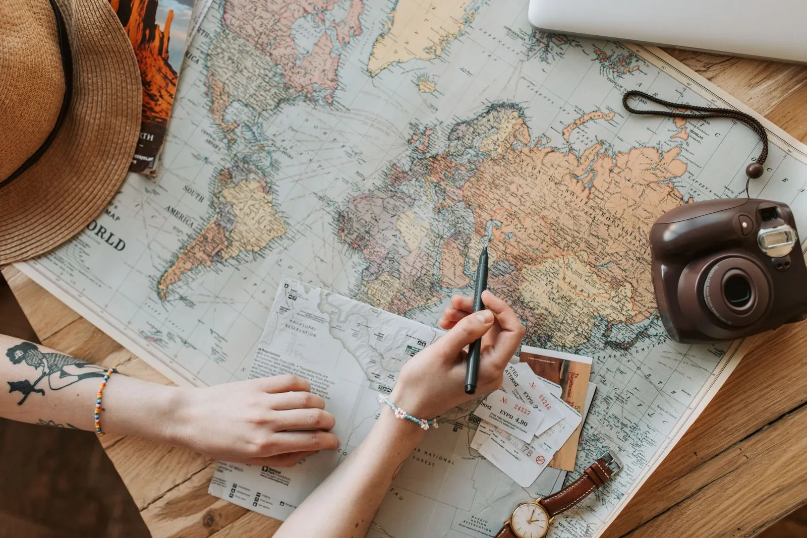 Hands planning a trip with a world map, a pen, tickets and an instant camera on a wooden table