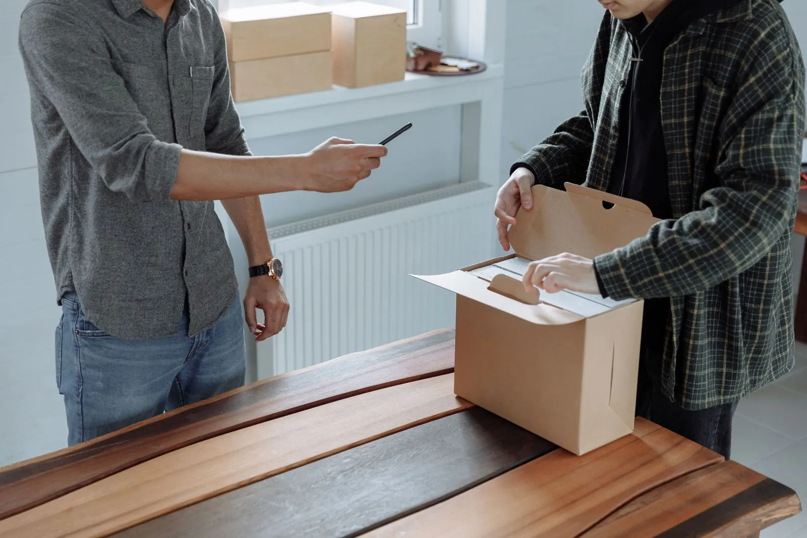 Two people opening a cardboard box while one of them holds a smartphone ready to take a photo
